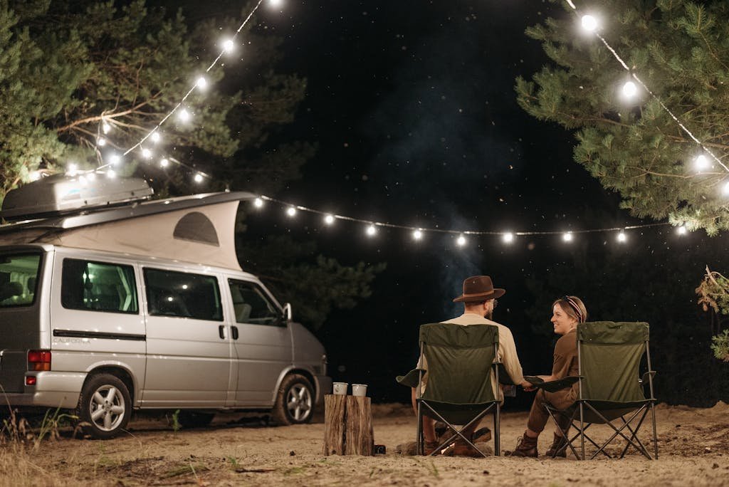 A couple sitting by their van under string lights, enjoying a quiet camping night.